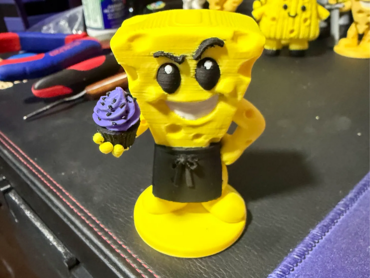 Cheezy Baker figure wearing a dark apron and holding a purple-frosted cupcake